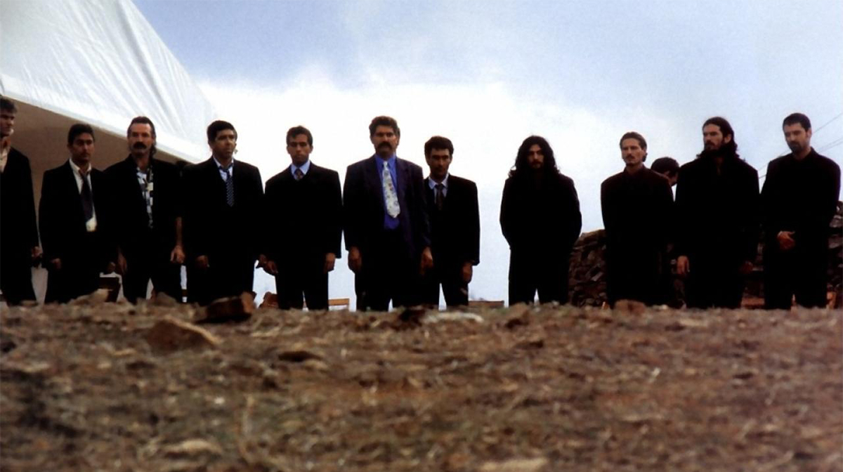 Eleven men in dark suits stand in a line and stare ahead at a pile of dirt